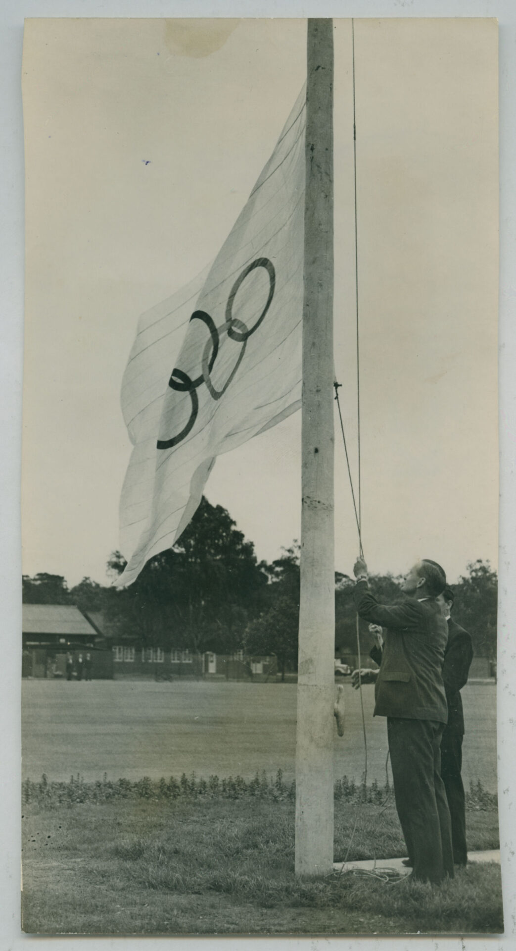 In a vintage black and white photo, a man displays the Olympic flag with its famous rings at the 1948 Olympic Village in Richmond Park.