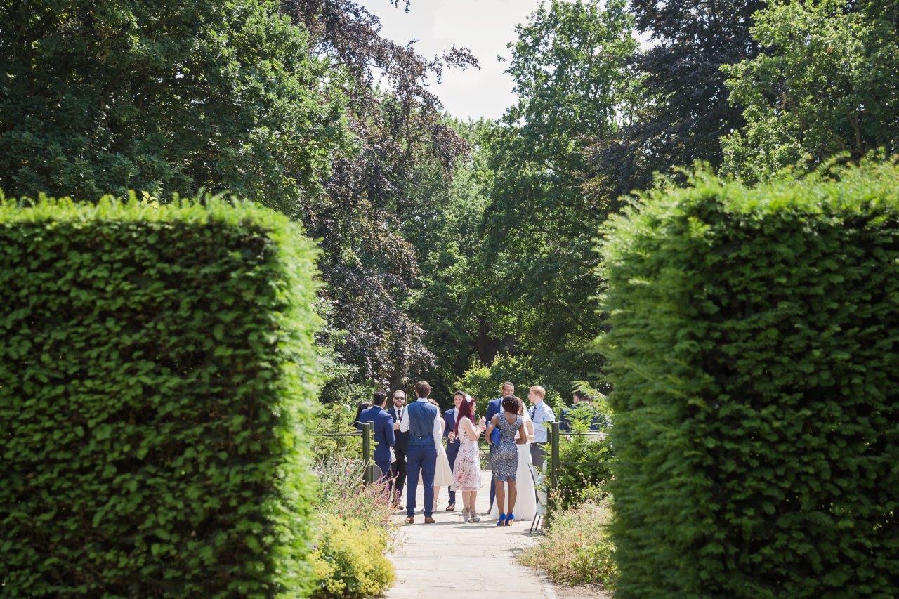 A view of wedding guests talking from a gap in a hedge.