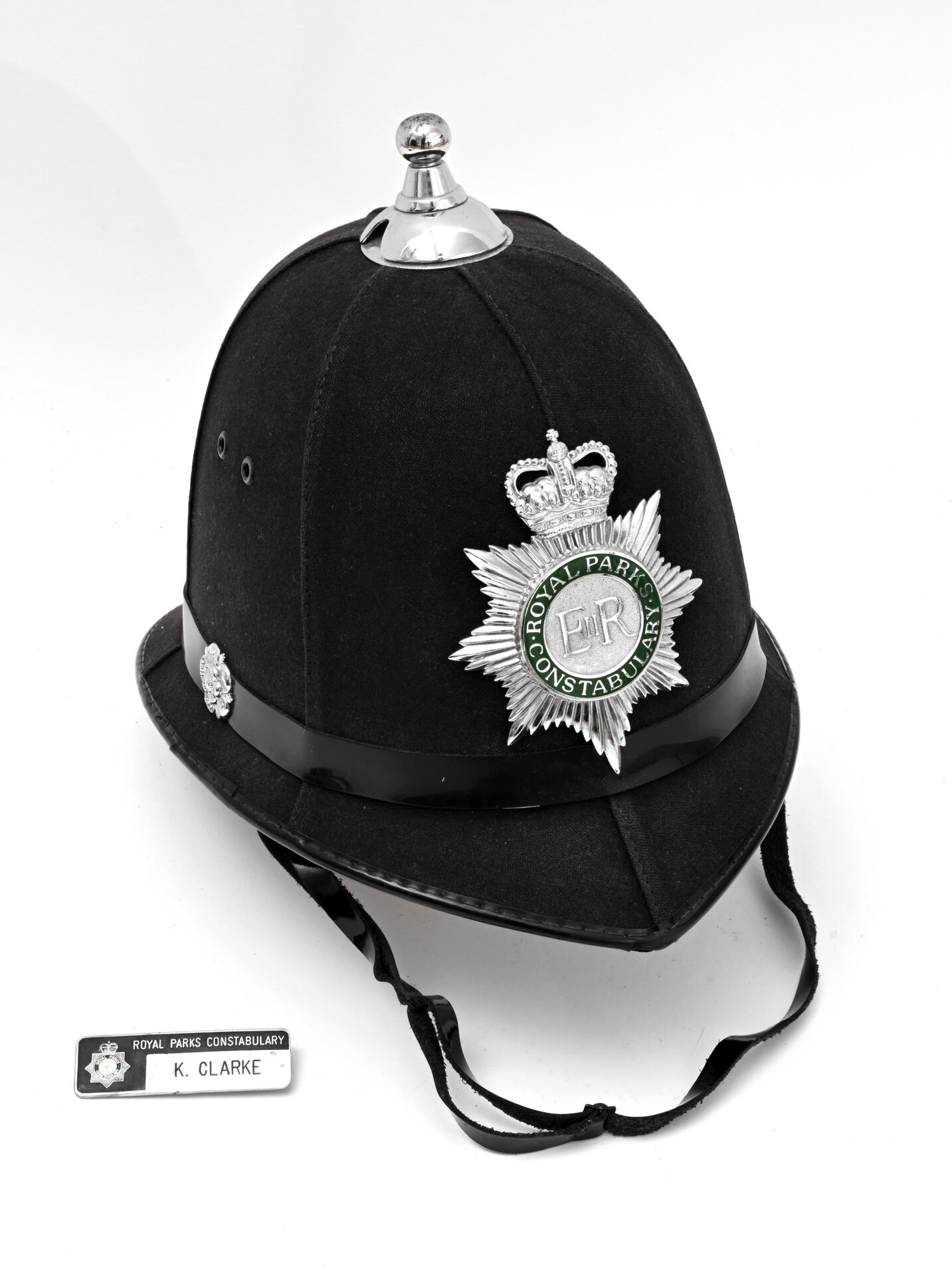 Black police helmet with a silver badge of the Royal Parks Constabulary. A name tag reading "K. Clarke" lies beside it. The tone is formal and historical.