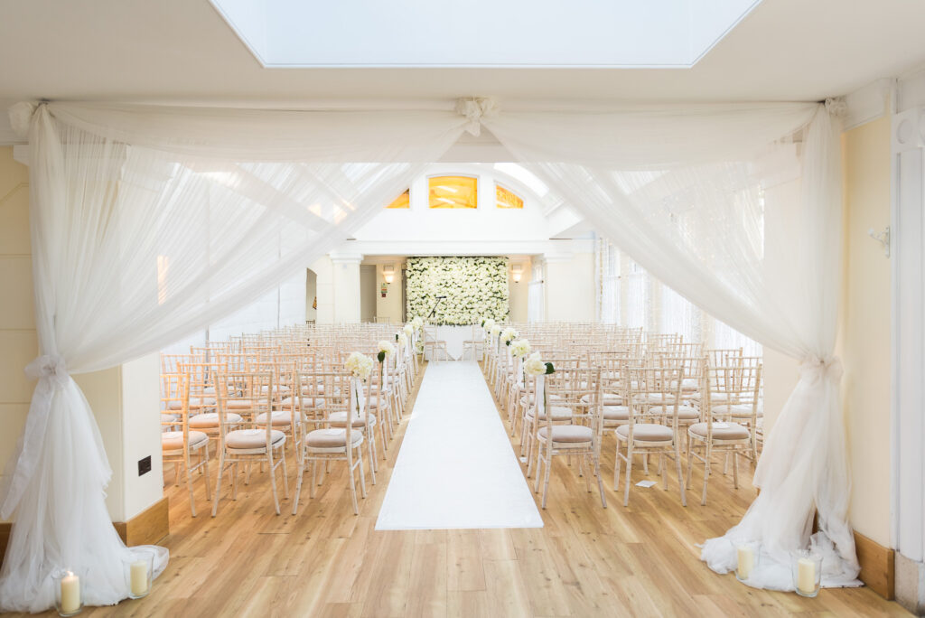Sheer white curtains drawn back to reveal a large room with white roses and rows of chairs.
