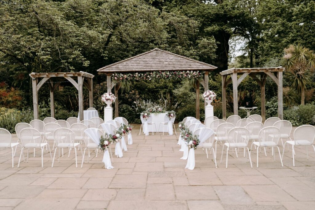 Outdoor wedding setup featuring rows of white chairs, floral arrangements, and a decorated arch in a lush garden.
