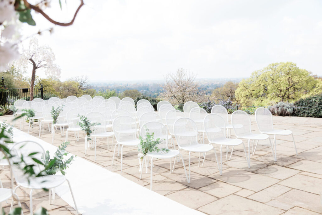Rows of white metal chairs with plants arranged on an outdoor patio.