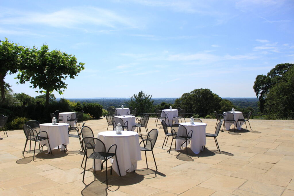 A large outdoor patio area with black metal chairs and round tables covered in white tablecloths.