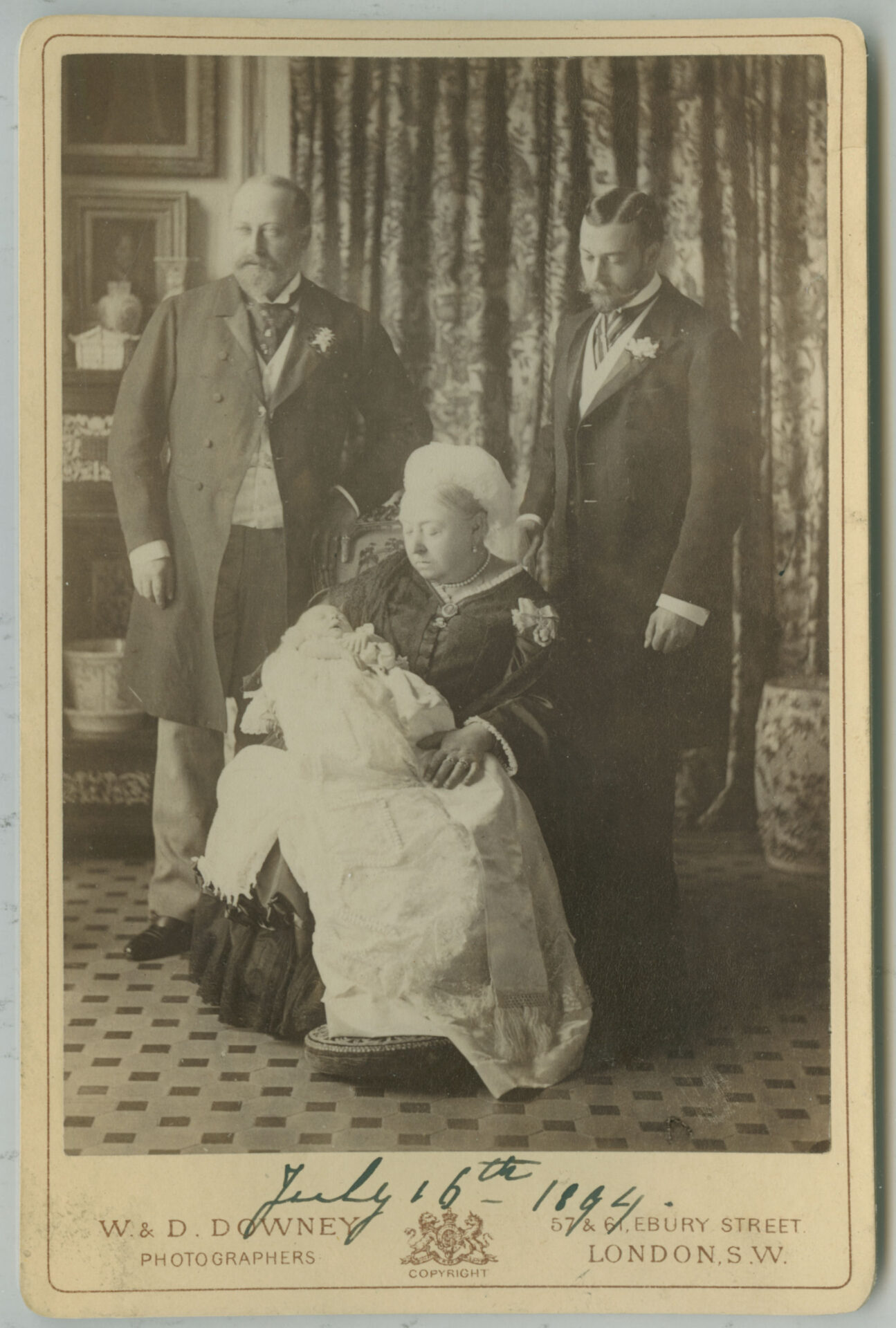 A historic photo of Queen Victoria holding baby Edward VIII, flanked by two formally dressed guests at the christening.
