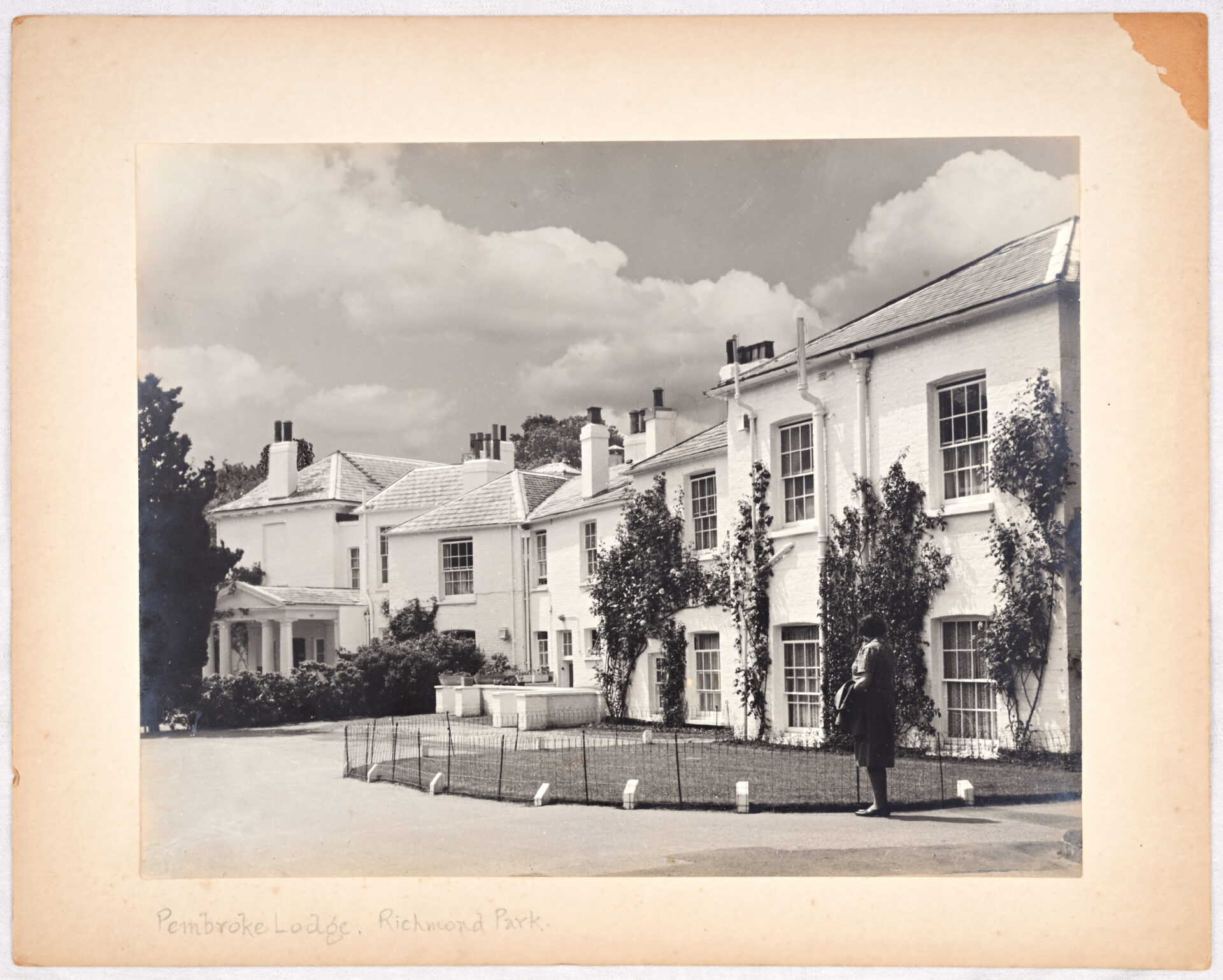 A woman strolls past a charming Pembroke Lodge featuring a bright white roof under a clear blue sky. Black and White Photo.