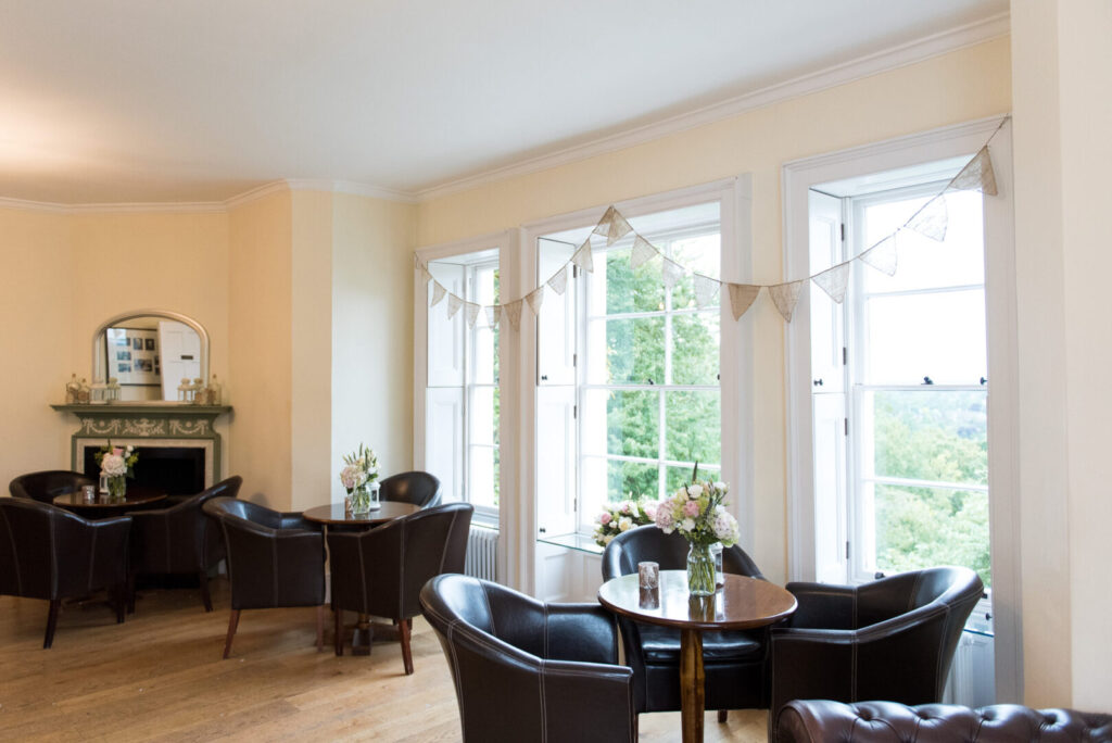 A large room with tall windows adorned by bunting featuring brown leather chairs and wooden tables