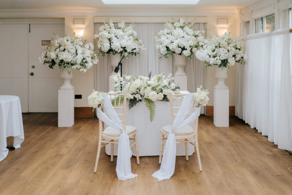 Two wooden chairs in front of a table covered in white cloth and flowers.