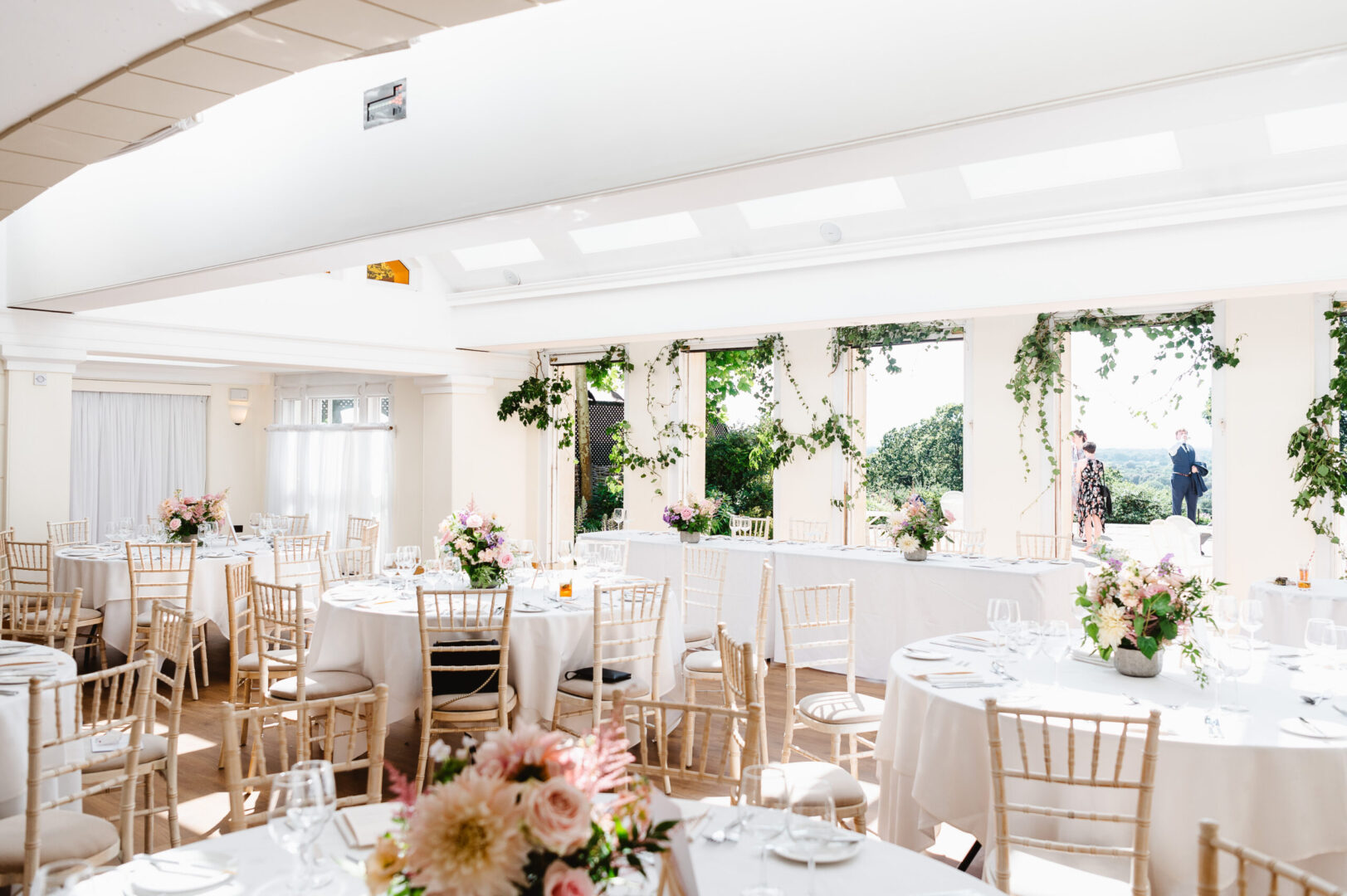 A bright room with open doorways and ivy, complimented by round tables with white tablecloths.