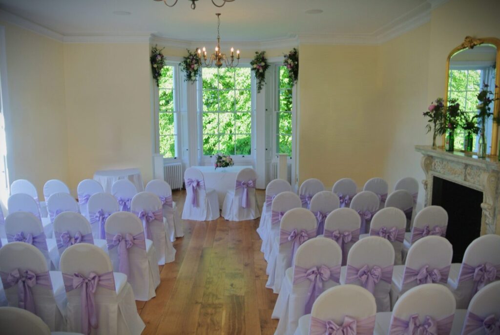 A large room with rows of white covered chairs with lilac bows.