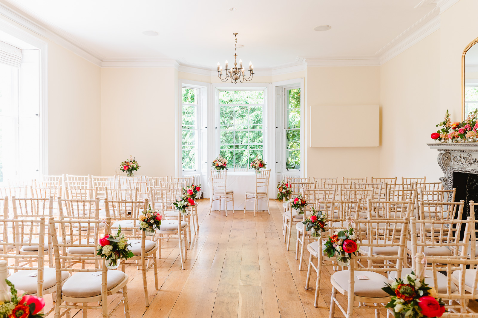 A bright room with large windows, rows of wooden chairs and bright floral arrangements.