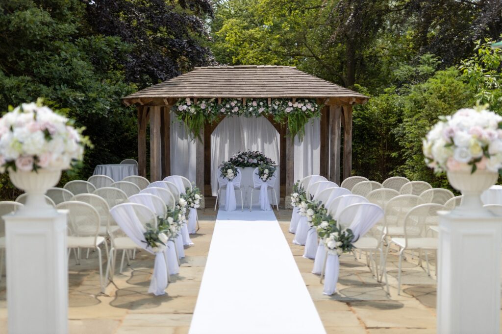 A wwedding arch with flowers and rows of white chairs in a patio area.
