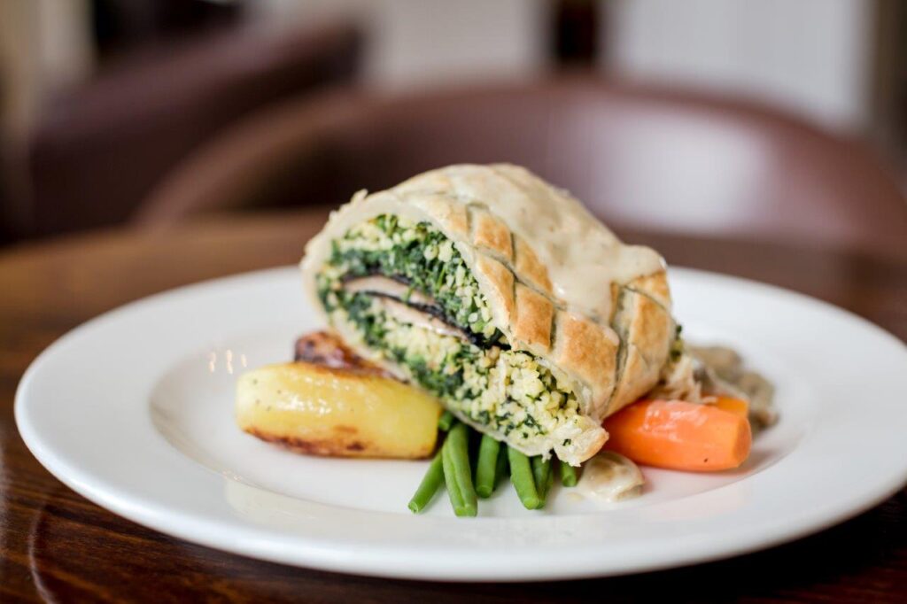 A vegetarian Wellington with green filling, surrounded by grilled potatoes, carrots, and beans on a white plate, conveying a warm, gourmet tone.