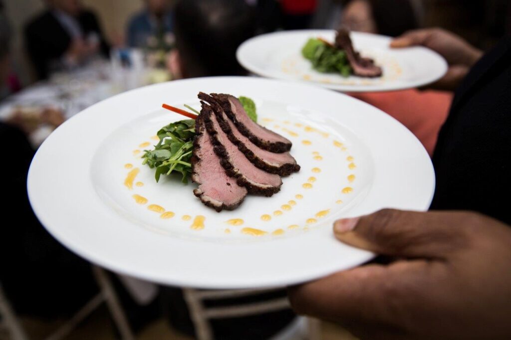 Server holding two plates of sliced, seared duck breast garnished with greens and sauce dots.