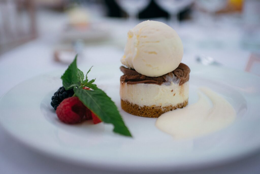 A cheesecake topped with ice cream on a white plate, garnished with mint leaves, berries, and a swirl of cream sauce; elegantly presented.