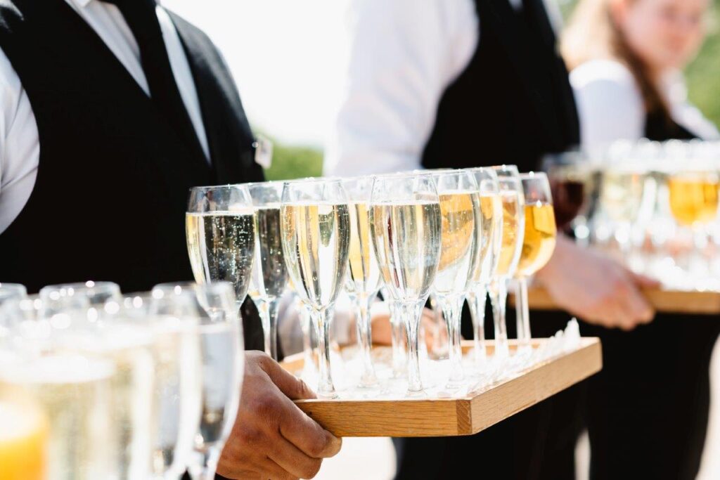 Servers in formal attire carry trays of champagne flutes at an outdoor event.
