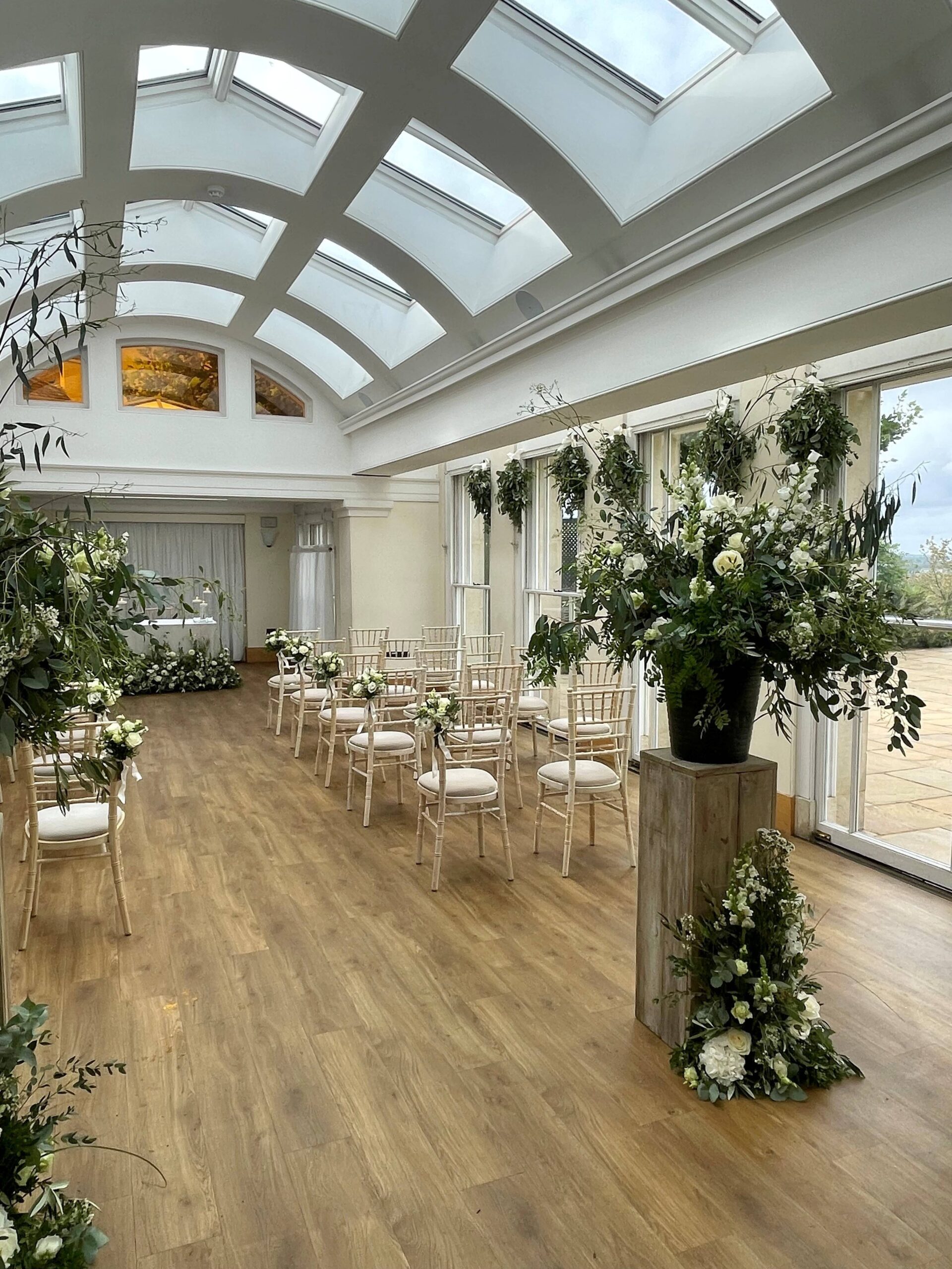 An ornate room with wooden flooring, rows of chairs and floral arrangements.