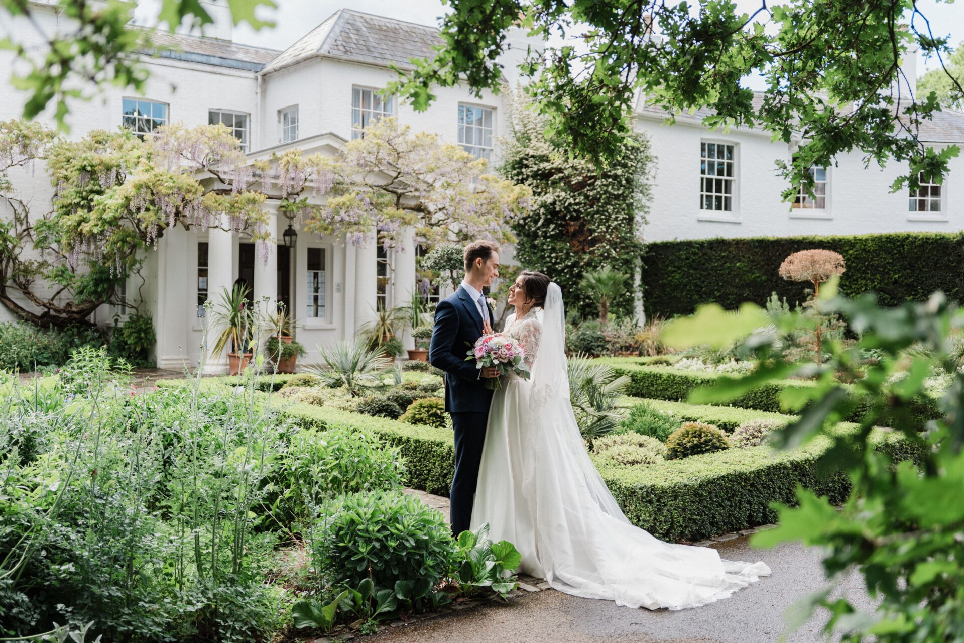 A bride and groom embrace in a large green landscaped garden.